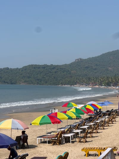 A scenic view of a beach with an adjacent mountain covered in lush greenery. The shoreline is lined with colorful umbrellas and beach chairs. Gentle waves touch the shore. The sky is clear and blue. A few individuals are visible on the beach, enjoying the scenery.