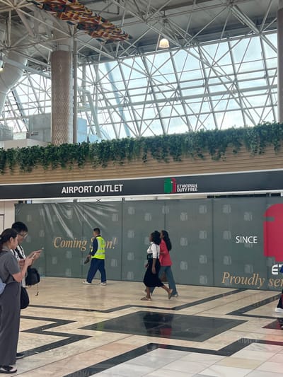 The image depicts an indoor scene at what appears to be Bole Addis Ababa International Airport in Addis Ababa, Ethiopia. The high ceiling is constructed of steel beams. On the right side of the image, text states, "Ethiopian Duty Free, Since", and "Proudly serving". The text "Airport Outlet" is also visible, and underneath that text is "Coming soon!". Several people are present, including two people sitting on the floor looking at their phones, three people walking in the background, and an airport employee in a yellow vest. The floor is light-colored, shiny tile with dark brown geometric patterns. Decorative green vines line the upper wall area.