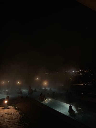 A nighttime scene depicts people bathing in an outdoor pool. Wisps of mist or steam rise from the water's surface, illuminated by submerged lights. The edges of the pool are visible, lined with snow. Figures are silhouetted against the dimly lit background, some standing near the pool's edge and others submerged. Above, faint building structures and lights are visible.