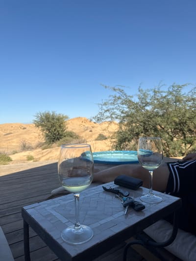 The image displays an outdoor leisure scene within the Merhavim Regional Council, Israel, under a clear, bright blue sky. In the foreground, a square table with a grey and white mosaic tiled surface supports two clear stemmed wine glasses. The left glass contains a small quantity of light-colored liquid, while the right glass is nearly empty. Positioned between the glasses are a closed black wallet, a silver and black corkscrew, and a set of vehicle keys featuring a black fob and a metallic key. Partially visible behind the table and glasses, a person is reclining on a wooden deck; only their lower torso and legs, clad in dark shorts with white horizontal stripes, are discernible. Beyond the reclining individual, a small, light blue inflatable pool is partially visible. The background consists of an arid, desert-like landscape with undulating light brown terrain and scattered green, shrub-like trees.