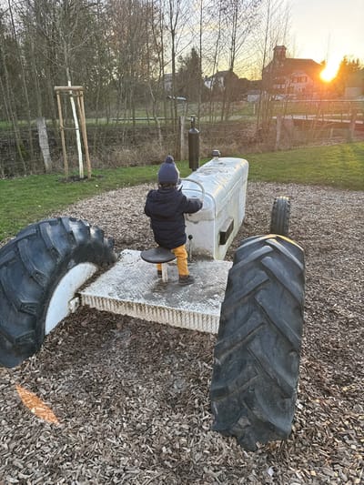 A young child is seated on a stationary playground structure resembling a tractor within an outdoor area in Olingen, Luxembourg. The child, viewed from the rear, wears a dark blue puffer jacket, yellow pants, dark shoes, and a dark blue knitted hat with a pompom, and is holding a white steering wheel. The tractor structure is primarily white, appears to be constructed from concrete or a similar material, and exhibits dirt and weathering. It features a black exhaust pipe and a black circular seat on which the child is positioned. Large, dark rubber tires are embedded into the wood chip ground around the structure, simulating tractor wheels. To the left, a grassy area borders a fence and numerous bare deciduous trees, with a young tree supported by wooden stakes. In the right background, a large building with a multi-gabled roofline is visible, partially silhouetted by the low-angle setting sun, which creates a bright orange flare. The ground surrounding the tractor is covered in light-colored wood chips.