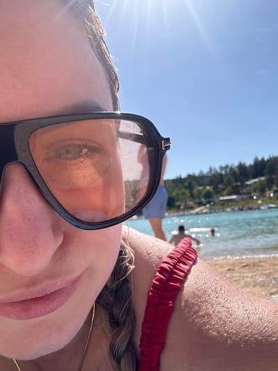 In Maisod, France, on a sunny day, a close-up shot focuses on the left side of a person's face, partially obscured by large black sunglasses with orange-tinted lenses. The person is wearing a red swimsuit strap across their shoulder. The background depicts a body of water, likely a lake, with people swimming and a shoreline lined with trees and buildings. The sky is a clear, bright blue with the sun glinting intensely, creating lens flare.