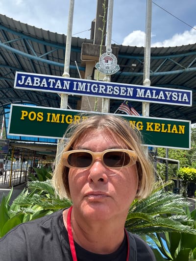 A selfie photograph shows a woman in sunglasses standing in front of the Malaysia immigration checkpoint in Wang Kelian, Malaysia. The "JABATAN IMIGRESEN MALAYSIA" sign is visible, along with another sign saying "POS IMIGRASI WANG KELIAN." A Malaysian flag is also visible. The environment includes vegetation and the structure of the immigration facility. The woman appears to be the sole subject of focus.