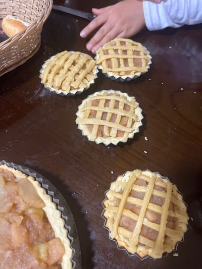 This image depicts a close-up, eye-level view of a dark wooden table on which several small, apple-filled tarts with lattice crusts are arranged. One larger tart with a similar apple filling is partially visible on the left side of the frame. A woven basket containing a bread roll is positioned in the upper left corner. A child's hand and arm, wearing a striped shirt, are visible in the upper right portion of the frame, reaching towards the tarts. The scene appears to be in a domestic setting, likely a kitchen or dining area. There is no discernible information about the specific location within San Luis, Peru, in this image.