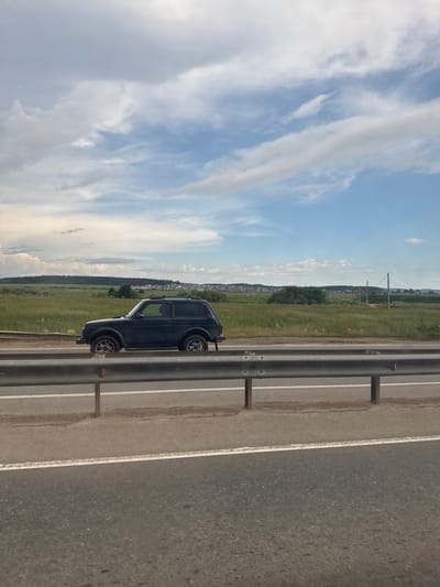 The image shows a dark-colored SUV, likely a Lada Niva, parked on the shoulder of a paved road in Kitsjanovo, Russia. The vehicle is positioned facing right in the frame, with its left side visible. The road has white lane markings and a metal guardrail runs along the right side of the frame. Beyond the road is a grassy field that stretches towards a treeline and a small village in the distance. The sky above is blue with scattered white clouds. The lighting suggests it is daytime, likely late afternoon or early evening, given the angle of the sun and the shadows. The weather appears to be fair. There is no text visible in the image.