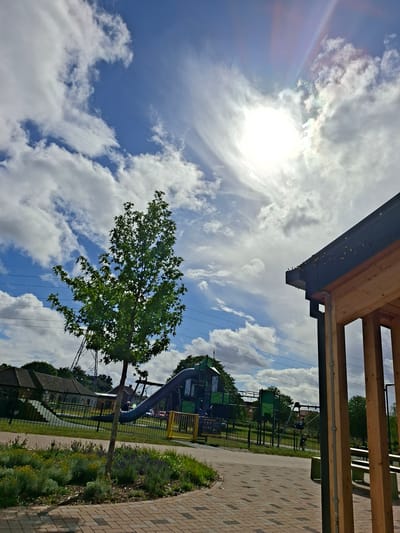 This image presents a bright, upward-angled view of an outdoor recreational area, likely a park or playground in Dartford, United Kingdom.

The sky is a prominent subject, displaying a vivid blue canvas dotted with large, fluffy white cumulus clouds. The sun is intensely bright, positioned high in the sky, causing significant lens flare and creating iridescent, rainbow-like effects within the clouds immediately surrounding it.

In the foreground, the underside of a modern structure, possibly a shelter or part of a building, is visible. It features exposed wooden beams and dark metal guttering, along which small, warm-toned lights, resembling fairy lights, are strung.

A deciduous tree with lush green foliage stands in the mid-ground, its branches reaching towards the sky. Beyond the tree, thin power lines or overhead cables are visible against the backdrop of the sky and clouds.

To the right, a light brown paved path made of interlocking bricks meanders through the scene. This path is bordered by a black metal fence, beyond which lies a children's playground. The playground equipment is colorful, primarily green and yellow, and includes slides and climbing structures. Surrounding the path and playground are patches of green grass and various shrubs and flowering plants.

No specific activity or individuals are discernible in the image, although the playground suggests recreational use. The scene is illuminated by strong sunlight, indicating it is daytime, likely mid-day, under clear and sunny weather conditions with scattered clouds. No legible text is visible on any of the objects.