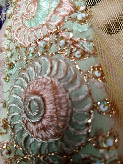 A close-up showcases intricate embroidery on a light green fabric. The design features spiraling shapes in light orange thread, embellished with metallic gold threads and small, iridescent beads. A sheer, golden mesh fabric appears in the background. The embroidery adds texture and visual interest to the soft fabric, creating a delicate and ornate pattern.