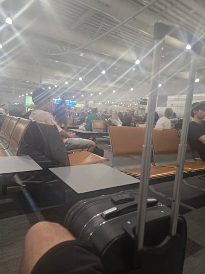 The image depicts the interior of an airport terminal, likely within the Spata-Loutsa Municipal Unit, Greece, characterized by its high ceilings, numerous light fixtures, and rows of seating. In the foreground, a dark-colored suitcase with a retractable handle is prominently positioned. A person's leg, wearing black shorts, is visible to the left of the suitcase.
The midground reveals multiple rows of tan-colored airport seating with metal frames, some occupied by individuals. A person in a light pink t-shirt and denim shorts is seated on the left, facing forward with their hands clasped. Behind them, other travelers are seated, some appearing to look at screens or converse.
The background shows more seating areas and a wall with what appear to be departure or information screens displaying digital text and graphics. The overall environment is brightly lit by overhead lights, with some glare visible, suggesting it is daytime. The architecture suggests a modern airport facility.