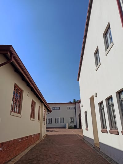 The image depicts a sunlit courtyard area with red brick paving. Two white buildings with red trim on their roofs are visible. The building on the left has a lower profile with several windows featuring dark frames and wooden mullions. Its lower section is clad in red brickwork. The building on the right is taller and more modern in style, also white with red trim, and features a series of vertically oriented windows with dark frames. In the background, another white building with a simple rectangular design and dark-framed windows can be seen. The sky is a clear, bright blue. The scene appears to be an outdoor educational or administrative campus setting, potentially within Pretoria, South Africa, given the architectural style and clear sky suggesting a warm climate. No people or animals are visible in the frame.