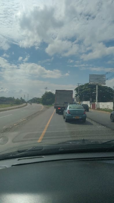 The image displays a daytime view from within a vehicle, looking forward through the windshield onto a multi-lane highway or main road. The sky above is blue with scattered white cumulus clouds, and bright sunlight is evident. The vehicle's dashboard and the base of the windshield are visible in the foreground.

On the asphalt roadway, a light blue sedan with a yellow Colombian license plate (partially visible as MGD-XXX) is positioned directly ahead. Behind the sedan is a dark grey box truck, displaying two illuminated red taillights. To the far right, a dark-colored vehicle is partially visible. Further down the road, to the left, the road curves, and a motorcycle is discernible in the distance, alongside other indistinct vehicles.

The environment to the right of the road features dense green foliage, a white concrete wall, utility poles supporting power lines, and a large, unadorned billboard structure. The left side of the road shows open land with sparse vegetation and faint, distant structures. No individuals are clearly visible. The vehicles appear to be in traffic. This scene is located on a main thoroughfare within or near Candelaria, Colombia.