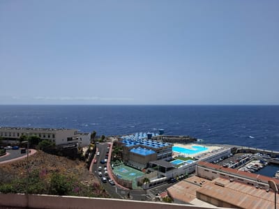 This is a high-angle, wide shot of a coastal area in Radazul Bajo, Spain. The scene is dominated by the deep blue ocean under a clear, pale blue sky. In the foreground, a white concrete wall and some green vegetation with pink flowers are visible.

The midground features a hillside with a winding road and several parked cars, including a black SUV, a blue sedan, and numerous white and silver cars. A large, modern white building with multiple windows is situated on the hillside overlooking the ocean.

Further into the scene, a complex of buildings with striking blue, angular roofs is prominent. Adjacent to this complex is a brightly lit blue swimming pool with lounge chairs. A smaller swimming pool is also visible nearby. A sports court, possibly for basketball, with colorful markings is situated in front of the blue-roofed buildings. A parking lot filled with cars and a marina with numerous boats docked are to the right of the sports court. Various other buildings, some with rusty, corrugated metal roofs, are scattered throughout the landscape. The overall impression is of a resort or residential area situated on a scenic coastline.