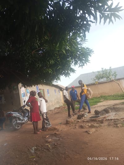 The image shows a street scene in Dokan Tofa, Nigeria. Several people are gathered, some appearing to be in conversation. A motorcycle is parked to the left, and the background consists of a building with posters on its exterior wall and a corrugated metal roof. The ground is dirt and appears to be somewhat muddy in places. The lighting suggests it is daytime, with the sun high in the sky.