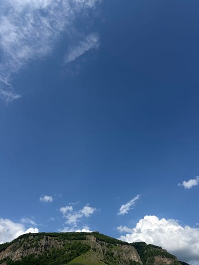 This is a landscape photograph featuring a vast expanse of a clear blue sky, punctuated by scattered white clouds. The right side of the image is dominated by a steep, rocky mountainside densely covered in lush green vegetation. The sunlight illuminates the clouds, making them appear bright and fluffy, while casting shadows on the mountain's textured surface.
The scene suggests a bright, sunny day with good visibility. The presence of the mountain and the clear sky implies an outdoor natural setting, likely in a mountainous or hilly region. The greenery on the mountain indicates a temperate climate, possibly during spring, summer, or early autumn.
The photograph is oriented vertically, with the sky occupying the majority of the frame and the mountain forming a dramatic diagonal line across the right side. There is no visible text in the image. The overall mood is serene and majestic, highlighting the beauty of nature. The specific location is Каменномостский, Russia.