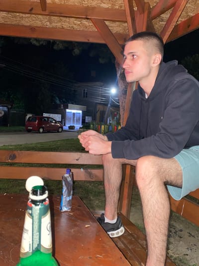 The image depicts a young man sitting on a wooden bench in what appears to be an outdoor seating area with a wooden roof structure. He is wearing a black hoodie and light blue shorts. His legs are bare and hairy. In the foreground, on a wooden table, there is a green bottle with a white label, a white cap on the bottle opening, and a silver pouch. Behind the bench, there is a grassy area, a street with a red car parked, and a building with illuminated windows. The scene appears to be at night, indicated by the dark sky and streetlights. The location is identified as Yasen, Bulgaria.
