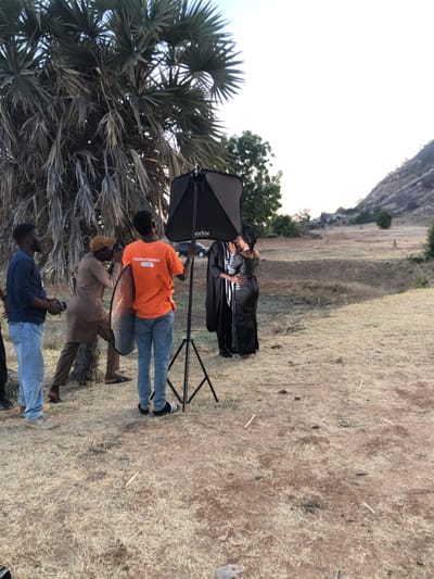 Outdoors, a woman in a long, black dress and striped top stands next to a man. A black Godox softbox on a stand is positioned to their left. Two men stand near them. One holds a reflector, wearing an orange shirt and blue jeans. Another man is nearby with a camera, wearing a tan shirt and tan pants. The sky is bright and the landscape is dry and grassy with a large tree to the left and a mountainous background.