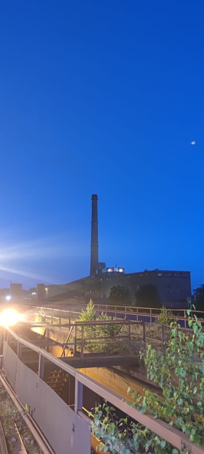 The image depicts an industrial landscape at dusk in Salzgitter, Germany. A tall, cylindrical smokestack dominates the skyline, emitting no visible smoke. Adjacent to it is a large, dark industrial building with illuminated windows. In the foreground, a weathered metal walkway with railings extends into the frame, bordered by sparse vegetation and some metallic structures. The sky is a deep, clear blue, transitioning to darker hues at the top, with a faint white object, possibly the moon or a distant light, visible in the upper right quadrant. The scene is illuminated by artificial lights, one of which is a bright, hazy orange light source on the left side of the image, casting a glow on the surrounding structures. The overall impression is one of an operational industrial facility under a twilight sky.
