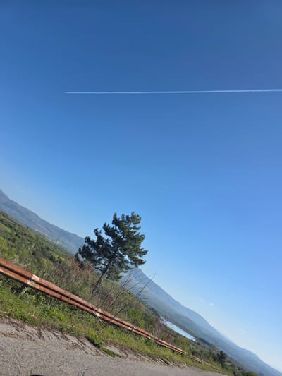 The image shows a scene in Lelintsi, Bulgaria. A blue sky with a contrail stretches above a landscape. The ground slopes downhill, with a road running along a ridge. A rusty metal guardrail separates the road from the edge. A single pine tree stands near the road, with hills and valleys visible in the distance. A body of water is also present in the distance. The photo appears to be taken during daylight hours.