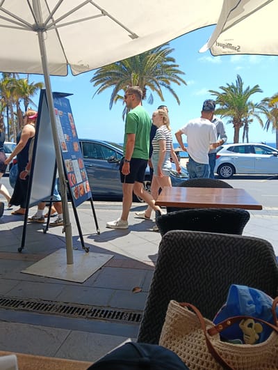 An outdoor scene in Puerto Del Carmen, Spain, captures a bustling sidewalk and cafe terrace under bright daylight. A large white patio umbrella dominates the foreground, offering shade, with the word "San Miguel" and "CERVEZAS SINCE 1890" visible in reverse on its underside.
On the sidewalk, several people are walking casually. A man in a green t-shirt and black shorts, wearing sunglasses, walks past an A-frame menu board that prominently displays "TAPAS" and various food items with accompanying images and prices. Behind him, a woman in a striped t-shirt and light shorts follows. Further along, another man in a white t-shirt and jeans is walking, while a woman in a dark dress and pink visor moves in the opposite direction.
The setting is distinctly coastal, with lush palm trees lining the street and the bright blue ocean visible in the background beneath a clear sky with scattered clouds. Cars are parked or driving on the road adjacent to the sidewalk.
In the immediate foreground, part of a cafe seating area is visible, featuring a wooden table and dark, wicker-patterned chairs. On one of the chairs, a light-colored crocheted bag or basket holds a blue item, possibly a jacket, and a small yellow toy, possibly a character like Mickey Mouse. The ground is paved with tiles, and a long metal grate runs along the edge of the sidewalk. The sunny weather and light clothing worn by pedestrians indicate a warm climate.