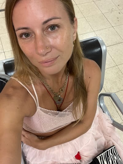 A woman with blonde hair, wearing a light pink lace bralette and a matching ruffled skirt, sits in a waiting area. She has a gold nose ring and multiple necklaces. Her arms are crossed over her chest. The background shows a tiled floor and a black and silver chair. The location is identified as Katunayake, Sri Lanka.