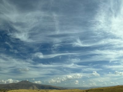 The image displays an expansive daytime landscape in Arzni, Armenia. The upper two-thirds of the frame are occupied by a deep blue sky, featuring numerous wispy white cirrus clouds stretching horizontally across the view and scattered cumulus clouds closer to the horizon. A faint white contrail from an aircraft is visible in the upper right quadrant of the sky. The lower third of the image shows an arid, undulating terrain covered in yellowish-brown dry vegetation. A prominent, dark-colored, conical mountain peak rises from the mid-ground on the left, with smaller, rolling hills extending towards the right horizon. No people or man-made structures are distinctly visible.