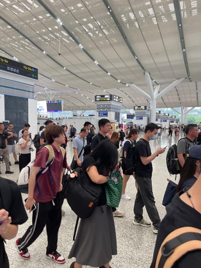 An overhead shot captures a bustling scene within a modern, well-lit transportation hub in Longhua District, China, likely an airport or train station. The spacious interior features a high, arched ceiling with a metal lattice structure and numerous recessed lights. White, tree-like columns support the roof. In the foreground, a diverse crowd of people, primarily appearing to be of East Asian descent, moves through the concourse. Many are carrying luggage, including backpacks and suitcases. Several individuals are looking at their mobile phones, while others are engaged in conversation or walking purposefully. Signage with Chinese characters is visible in the background, indicating different gate or platform numbers. The floor is a light-colored speckled material. The overall impression is one of travel and transit in a contemporary urban environment.