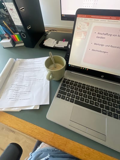 The image shows a desk with a laptop, papers, and a mug of coffee. The laptop screen displays a presentation slide with bullet points in German, including "Anschaffung von Maschinen und Geräten" (Acquisition of machinery and equipment) and "Wartungs- und Reparaturkosten" (Maintenance and repair costs). A document with similar content is open on the desk. The environment appears to be an office or study area. The scene is likely located in Morsbach, Germany, based on the provided information. There are no people clearly visible in the image.