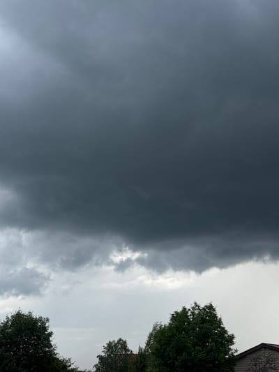 The image depicts a view of the sky from the city of Taskino, Russia. The sky is dominated by dark, ominous storm clouds that fill the upper portion of the frame. Below the storm clouds, lighter, more diffused clouds are visible, with hints of a brighter sky peeking through. The lower edge of the image shows the tops of several lush green trees and a portion of a building with a reddish-brown roof. The overall scene suggests an impending storm.
