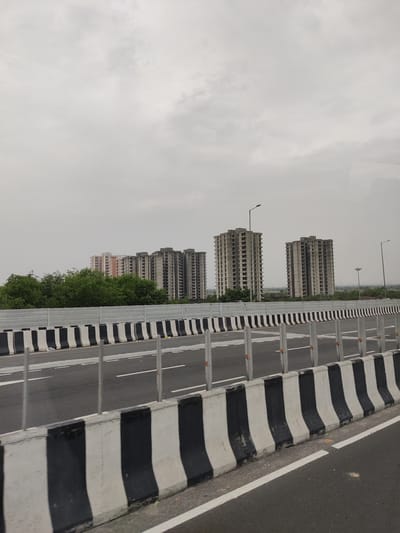 An overcast day in Ghaziabad, India. The image, captured from a moving vehicle on a highway, shows several tall, unfinished apartment buildings in the distance. These concrete structures have numerous open windows and appear to be under construction or abandoned. In the foreground, a highway barrier with alternating black and white painted sections lines the road. The road itself has white lane markings. Lush green trees are visible behind the barrier, partially obscuring some of the lower portions of the buildings. The sky is a uniform gray, indicating cloudy weather.