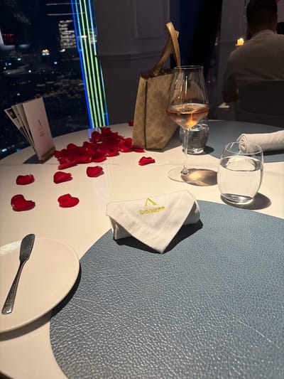 An indoor scene shows a table set for a romantic dinner with a cityscape visible through a window. The table has a white tablecloth and is adorned with red rose petals scattered around. On the table are a wine glass filled with rose-colored liquid, a glass of water, a white folded napkin with golden embroidery of a triangle and cursive text, and a small white plate with a silver butter knife. A beige handbag with a repeating logo pattern rests near the wine glass. In the background, a person is seated at another table, partially obscured. The window reveals a nighttime view of a city, with illuminated buildings and lights. A tall, modern structure with vertical green and blue lights is prominent in the cityscape. The overall ambiance suggests a celebratory or romantic dining experience. The inscription on the napkin appears to read "L'AROMATIQUE".