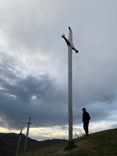 A low-angle shot taken at an odd, slightly tilted angle shows a figure standing beside a large, metal sword monument, with two other similar but smaller monuments in the background. The location is Zemo Nichbisi, Georgia. The sky is cloudy, and the ground is covered with grass. The person is wearing dark clothing and is positioned near the top of a small hill.