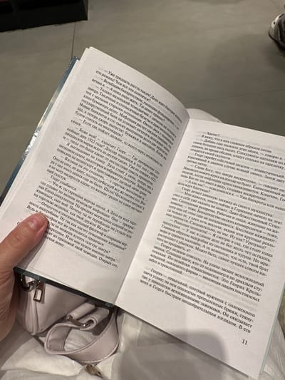 The image shows an open book with text on both pages, held by a person's hand with a visible thumb. The text discusses financial matters and a historical context, mentioning a "bankrupt," "inflation," and "bills." The right page contains the number "11" at the bottom. Below the book, a light-colored handbag with metal clasps and straps is visible. The setting appears to be indoors, with soft lighting. The city of Kozulino, Russia, is not mentioned or depicted in the image.