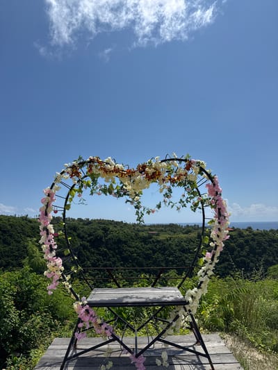 The image captures a heart-shaped arch decorated with pink, white, and orange flowers. The arch is positioned on a wooden platform overlooking a dense green forest. In the background, a body of water can be seen separating the forest from the sky. The sky is bright blue with some scattered clouds. The location is Pejukutan, Indonesia.