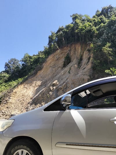 A silver car sits against a backdrop of a steep, eroded hillside, topped with dense green trees. The earth is a mix of pale browns and tans, showing signs of past landslides. The blue sky is visible above the trees and to the side of the hillside. The car’s tire, headlight, mirror, door, and tinted window are visible in detail.