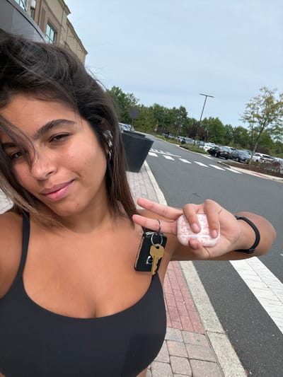 A female subject is depicted from the upper torso up, standing outdoors in Plainsboro Township, United States. She possesses dark hair, brown eyes, and a tanned complexion, engaging directly with the camera with a subtle smile. She wears a black athletic top and multiple silver hoop earrings in her left ear. Her left hand is raised, forming a peace sign, from which a black car key fob and a single brass key are suspended on a metal ring. Her right hand grasps a light pink AirPods case featuring a subtle patterned design. A black elastic wristband is present on her right wrist. The background features a paved sidewalk, an asphalt road with white pedestrian crossing markings, and a parking lot containing numerous vehicles. A line of green trees stands behind the parking area, beneath an overcast sky. A large, light-colored commercial or retail building is partially visible to the upper left, alongside a dark tall pole and a dark planter on the sidewalk.