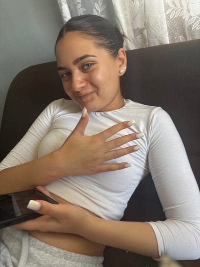 A young woman with dark hair pulled back sits on a dark brown couch. She is wearing a white long-sleeved shirt and grey sweatpants. Her left hand is resting on her chest, and her right hand holds a black smartphone. She has light-colored acrylic nails. The background includes a white lace curtain. There are no specific landmarks in Radievo, Bulgaria, visible in this image.