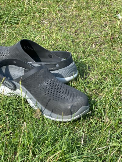 A pair of black and grey Crocs Literide Clogs rests on a patch of green grass under direct sunlight. The shoes, with their perforated uppers and textured grey soles, are positioned in the foreground, one slightly behind and to the right of the other. The grass appears natural and somewhat dry, with varying blade heights. No individuals are present. The scene is outdoors, likely in Nieporęt, Poland, during daytime with clear weather conditions.