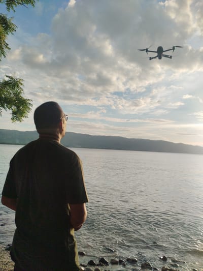 A medium shot depicts a man viewed from the back at the shore of a body of water, with a drone in flight in the sky. The man is wearing a dark t-shirt and glasses. The backdrop features a coastline with rolling hills, a wide expanse of water, and a sky filled with clouds. The drone is positioned above the man in the sky. The foreground shows a pebbly shoreline. The photo appears to be taken in Lamreh, Indonesia, based on the landscape and cultural references.