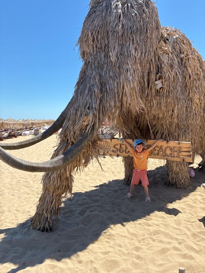 A young child stands with arms outstretched, leaning on a wooden sign that reads "SEA BEACH" in carved lettering. The sign is propped between two large, straw-covered columns that resemble the legs of a mammoth. The child is wearing a blue hat, an orange t-shirt, and coral-colored shorts. They are standing on a sandy beach, with rows of beach chairs and umbrellas visible in the background. The sky is clear and blue. The scene appears to be at a beach resort in Shkorpilovtsi, Bulgaria.