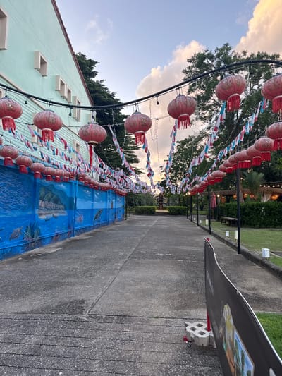 A pathway lined with red lanterns and small triangular flags leads towards a cluster of open-air huts at dusk in Ban Bang Niang, Thailand. The pathway is flanked by a turquoise building on the left, adorned with decorative beige window frames, and a vibrant blue mural depicting marine life and a village scene on the lower portion of the building's exterior wall. The mural features turtles, fish, dolphins, and a tropical island landscape. On the right, a manicured lawn borders the pathway, with a wooden bench, a closed red umbrella, and several small white lanterns placed along the edge. In the distance, lush green trees form a backdrop against a sky filled with pale clouds tinged with orange and pink from the setting sun. A large black banner on a red stand with a wheel is positioned in the foreground on the right, partially obscuring the view of the pathway. The pathway itself is made of concrete with visible cracks and lines.