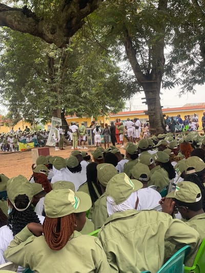 The image depicts an outdoor gathering of young adults, many wearing matching olive green uniforms and caps, at a facility with yellow buildings in the background. A large tree with a thick trunk and branches is a prominent feature, providing shade over a portion of the scene. The ground appears to be dirt. In the foreground, several individuals are seated, facing away from the camera, their heads and shoulders visible. They are wearing the aforementioned green uniforms and caps. In the mid-ground, a group of people are standing and gathered around a large poster displayed on an easel. Some individuals are dressed in similar green uniforms, while others wear white shirts and shorts, or other civilian clothing. Several people are seated on chairs in this area as well. The overall atmosphere suggests a formal event or assembly, possibly an orientation or ceremony, given the uniformed attendees. The location is identified as Emure-Ekiti, Nigeria.