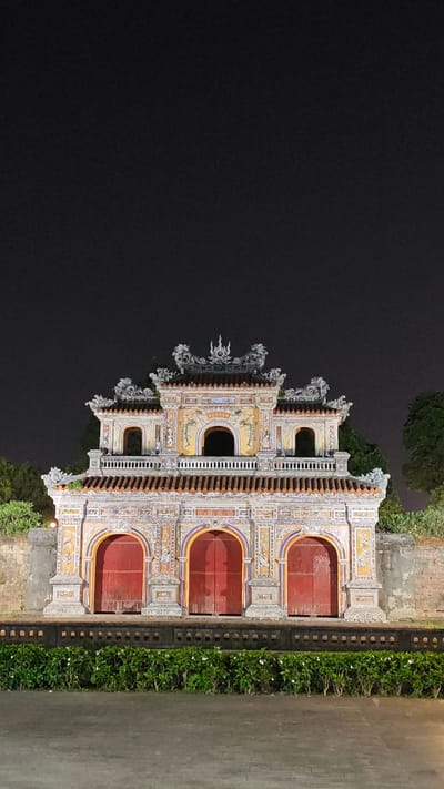 The image shows a brightly decorated three-arched gate at night. The gate's arches are framed by intricate designs in yellow, white, and blue. The doors behind the arches are painted red. Above the arches, there is a balcony-like structure with a balustrade. The upper section of the gate features more ornate designs and a tiled roof with decorative figurines. The sky behind the gate is dark, suggesting it is nighttime. Bushes with white flowers are planted in front of the gate. The location is Phú Xuân Ward, Vietnam.
