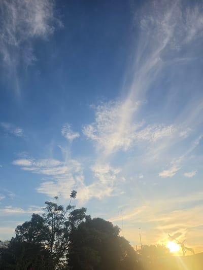 The image depicts a daytime sky with scattered clouds and a view of trees to the right. The sky ranges from deep blue in the lower left to a lighter, brighter blue mixed with white clouds towards the upper right where the sun is located. The sun's glare obscures specific details but suggests it's shining brightly.
Along the right edge of the image, dark tree foliage is visible, extending from the lower right corner upwards. At least one antenna is placed over the top of trees. The location is likely Pintomakmur, Indonesia. No people or specific actions are visible in the photograph.