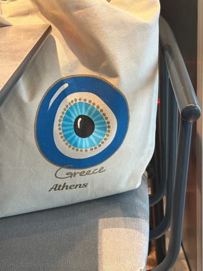 The image shows a cream-colored tote bag with a blue and black evil eye symbol printed on it. The words "Greece Athens" are printed in brown script to the right of the evil eye. The bag is draped over a gray metal chair. The location, Spata-Loutsa Municipal Unit, Greece, is indicated by the text on the bag.