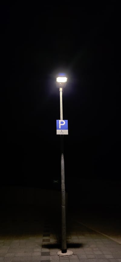 A single, illuminated street lamp stands against a black, night sky. The lamp is mounted on a grey metal pole that tapers towards the base. Attached to the pole, below the lamp, is a rectangular sign with a blue square background and a white "P" inside. Below the "P", there is a white symbol of a person in a wheelchair. The base of the lamp pole is secured by a concrete foundation. The ground beneath the lamp is paved with interlocking rectangular tiles, some of which are illuminated by the lamp's light, casting a shadow. The scene appears to be a pedestrian area or sidewalk in the city of Lieli, Switzerland, as indicated by the parking sign.