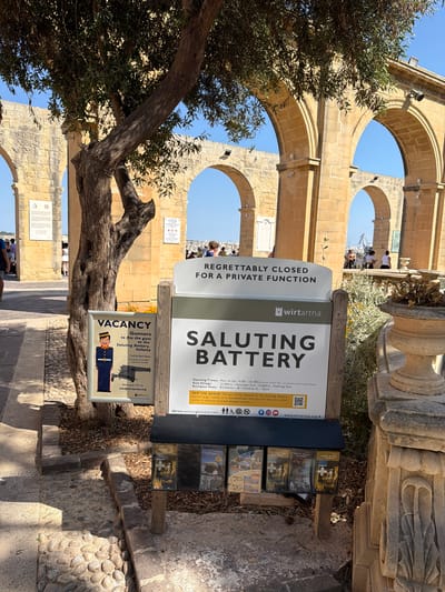 In Valletta, Malta, a tree with rough bark and sparse foliage stands in front of a stone structure with multiple arches. To the right of the tree, a sign announces that the "Saluting Battery" is regrettably closed for a private function. The sign includes operating hours, entrance fees, and a website address. Below this main sign, a smaller rack displays several brochures. To the left of the tree, another sign advertises a vacancy for "Gunners to fire the guns at the Saluting Battery, Valletta," with details on how to apply. In the background, people can be seen walking and observing the surroundings. The ground is covered with cobblestones and gravel. The sky is clear and blue.