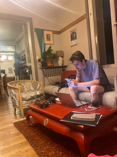 A person is seated on a grey couch while using a mobile phone and a laptop on a red table. There is a tan and white chair beside the red table and an oriental rug on the floor. The location appears to be an apartment, based on the interior details, but the specific location in Nanterre, France, cannot be determined from the visual information. Other items visible include framed art on the walls, potted plants, and headphones. The environment is lit with natural and artificial light.