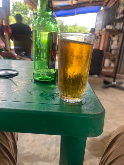 A green plastic table holds a glass of beer and a green beer bottle. There's condensation on the glass. Sunglasses are on the table as well. Part of a person's leg is visible next to the table. The background suggests an outdoor setting, possibly in Osogbo, Nigeria.