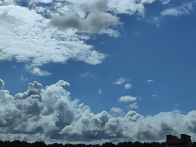 The image shows a wide, bright blue sky filled with dynamic white and grey cumulus clouds. The lower edge of the frame is lined with a dark silhouette of trees, indicating a terrestrial landscape. In the bottom right corner, a portion of a dark structure, possibly a building or a rooftop, is visible with a hint of red. The overall scene depicts a partly cloudy day, likely during daylight hours, over an area that includes natural vegetation and man-made structures. The location is specified as Salacgrīva, Latvia. There are no people or discernible actions depicted in the image.