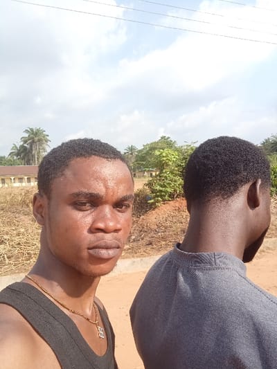 The image depicts two males outdoors in Omicha Umu Awada, Nigeria, under a partially cloudy sky. In the foreground, positioned on the left, an individual with dark skin, short dark curly hair, and a neutral expression faces the camera directly. He is wearing a black sleeveless top and a gold-colored chain with a pendant. His skin exhibits a sheen consistent with perspiration or oil. To his right, partially obscured, is a second individual with dark skin and short dark curly hair, seen from behind, facing away from the camera. This individual is wearing a grey long-sleeved shirt with a stand-up collar. The background features an unpaved reddish-brown ground surface. To the left, a mound of dry straw-like vegetation is present. Further back, various green trees and bushes are visible, along with multiple tall palm trees. A light-colored, single-story building with a brown roof is visible in the mid-left background. Overhead utility wires traverse the upper portion of the frame against the sky.
