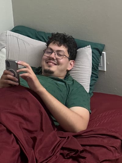 In an indoor setting, likely a bedroom in Franklin Township, United States of America, a young man is depicted lying in bed. He appears to be in his late teens or early twenties, with dark curly hair, glasses, and a short beard or goatee. He is wearing a dark green short-sleeved shirt. The man is lying on a light grey pillow, with a darker green pillow visible behind it, and is partially covered by a dark red or maroon blanket/comforter.
He is holding a dark gray smartphone in his right hand, with a circular attachment visible on its back, and appears to be interacting with the device. His face shows a broad, genuine smile, indicating a cheerful and relaxed mood, possibly amused by content on his phone. In the background, a plain light grey wall is visible, along with a white North American-style wall outlet with two sockets. The lighting suggests an indoor environment, but the specific time of day is not discernible. No visible text is present in the image.